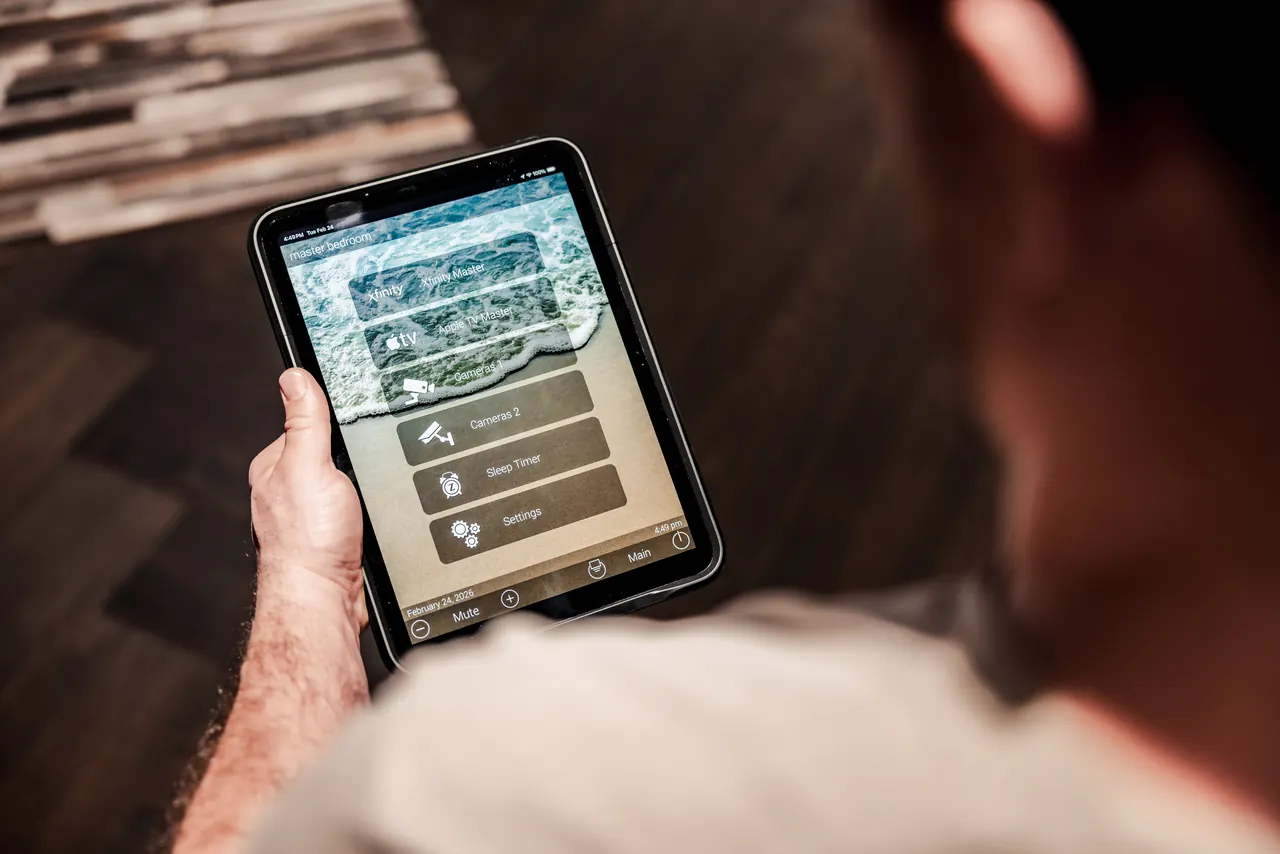 A homeowner holding a tablet running a custom home control interface with named sources and cameras
